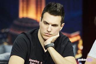 A professional poker player sits at the table with a focused expression, resting his chin on his hand. He is wearing a black Upswing Poker shirt, with a blurred background suggesting a televised poker event.